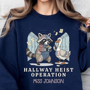 May include: Navy blue sweatshirt featuring a cartoon raccoon and goose with the text "Hallway Heist Operation Miss Johnson." The raccoon and goose are in front of a school locker background. The sweatshirt is a crew neck.