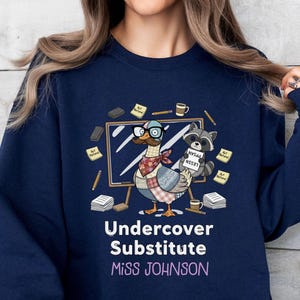 May include: Navy blue sweatshirt with a whimsical graphic of a goose and raccoon as a teacher. The goose wears glasses and a scarf, and the raccoon holds a sign. Text reads "Undercover Substitute Miss Johnson."