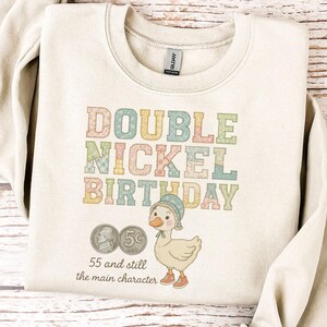 May include: Cream-colored sweatshirt with the text "DOUBLE NICKEL BIRTHDAY" in pastel colors. The design includes a cartoon duck wearing a hat and shoes, along with two nickels and the phrase "55 and still the main character."