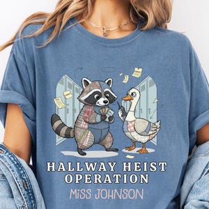 May include: Blue t-shirt with the text "HALLWAY HEIST OPERATION Miss JOHNSON". The graphic features a raccoon and a goose in front of lockers. The raccoon is wearing a patchwork outfit and the goose is wearing a patchwork outfit.