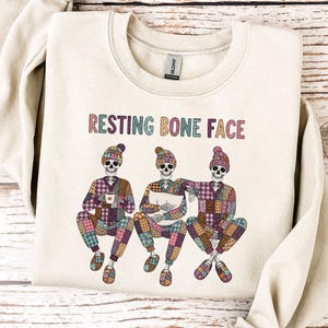 May include: Cream-colored sweatshirt with the phrase "RESTING BONE FACE" in colorful letters. The graphic features three cartoon skeletons wearing hats and patchwork outfits. One skeleton holds a cup, another a book.