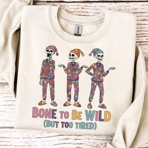 May include: Cream-colored sweatshirt featuring a graphic of three skeletons in colorful pajamas and hats. The text "BONE TO BE WILD (BUT TOO TIRED)" is printed below the skeletons in a retro font. The sweatshirt is laid on a wooden surface.