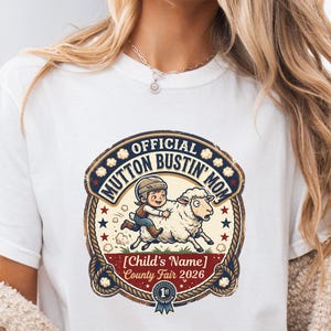 May include: A white t-shirt featuring a circular graphic with a rope border. The design shows a child riding a sheep with the text OFFICIAL MUTTON BUSTIN' MOM, [Child's Name], and County Fair 2026. A blue ribbon icon with 1st is at the bottom.