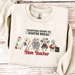 May include: Cream-colored sweatshirt with the text "Counting Down to Winter Break" and "Stem Teacher." The design features cartoon geese dressed for winter, holding items like an apple, laptop, and coffee. The sweatshirt is made of a soft material.