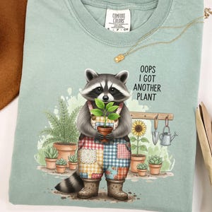 May include: A light green t-shirt featuring a raccoon wearing patchwork overalls and holding a potted plant. The text "Oops I Got Another Plant" is printed on the shirt. Other plants and gardening tools are also visible.