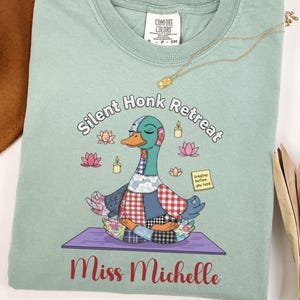 May include: A light green Comfort Colors t-shirt featuring a cartoon duck in a yoga pose. The shirt has the text "Silent Honk Retreat" and "Miss Michelle" printed on it. A gold necklace is draped on the shirt.