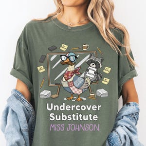 May include: Olive green t-shirt featuring a cartoon duck wearing glasses and a raccoon holding a sign. The shirt reads "Undercover Substitute Miss Johnson" in white and purple text. Pencils, coffee cups, and sticky notes surround the duck and raccoon.