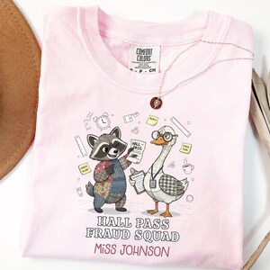 May include: A light pink Comfort Colors t-shirt featuring a cartoon raccoon and goose holding hall passes. The text "HALL PASS FRAUD SQUAD" and "Miss Johnson" are printed below the cartoon characters. A gold necklace is draped on the shirt.