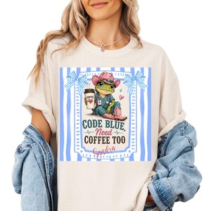 May include: Cream-colored t-shirt featuring a cartoon frog in scrubs and a cowboy hat, holding a coffee cup. The shirt has the text "CODE BLUE, NEED COFFEE TOO" and the name "Sandra" in a blue and white frame.