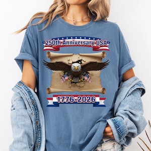May include: Blue t-shirt with a patriotic design. The shirt features an eagle with outstretched wings, holding American flags, and the text "250th Anniversary USA" and "1776-2026" in red, white, and blue.