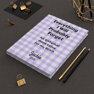 May include: A hardcover journal with a light purple and white gingham pattern. The cover reads "Everything I Will Probably Forget, An External Hard Drive for my Brain, Jackie 2026." Gold and black pencils and gold binder clips are nearby.