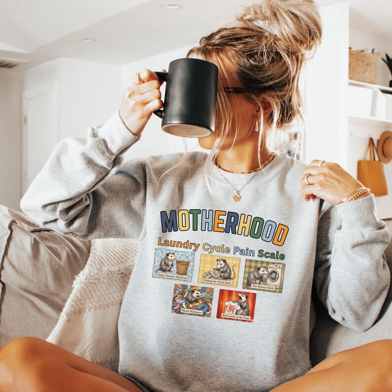 May include: A light gray sweatshirt with the word "MOTHERHOOD" in colorful letters. Below the word is "Laundry Cycle Pain Scale" and images of opossums. The person in the image is holding a black mug.