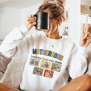 May include: White sweatshirt with the word "MOTHERHOOD" in colorful block letters. Below the word is "Laundry Cycle Pain Scale" and images of opossums. The person is holding a black mug.