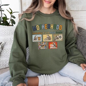 May include: Green sweatshirt with the word "MOTHERHOOD" in colorful letters. Below the word is "Laundry Cycle Pain Scale" and images of opossums. The sweatshirt is a solid green color.