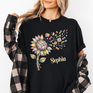 May include: Black t-shirt featuring a colorful sunflower design made of sewing notions. The sunflower includes spools of thread, scissors, and a measuring tape. The name "Sophia" is printed in a cursive font below the sunflower.