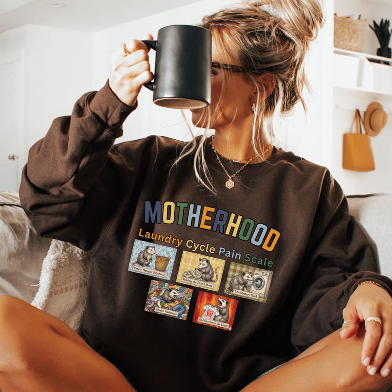 May include: A brown sweatshirt with the word "MOTHERHOOD" in colorful letters. Below the word is "Laundry Cycle Pain Scale" and images of opossums. The person in the image is holding a black mug.