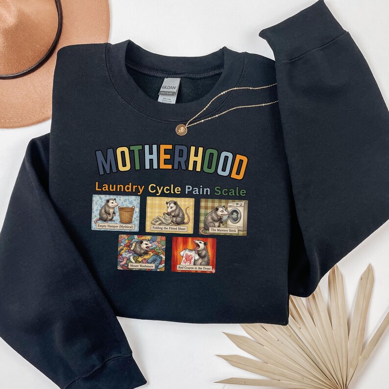 May include: Black sweatshirt with the word "MOTHERHOOD" in colorful letters. Below the word is "Laundry Cycle Pain Scale" and images of opossums doing laundry. A gold necklace and a tan hat are also in the image.
