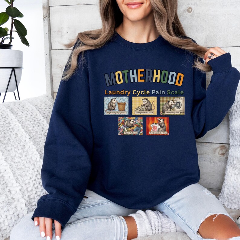 May include: Navy blue sweatshirt with the word "MOTHERHOOD" in colorful letters. Below the word is "Laundry Cycle Pain Scale" and a graphic of opossums doing laundry. The sweatshirt is a crew neck style.