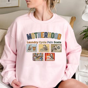 May include: A light pink sweatshirt with the word "MOTHERHOOD" in colorful block letters. Below the word is "Laundry Cycle Pain Scale" and six images of opossums doing laundry. The sweatshirt is made of a soft material.