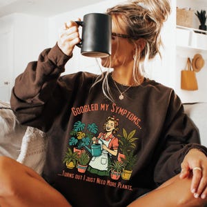 May include: A dark brown sweatshirt with the text "Googled My Symptoms... Turns Out I Just Need More Plants." The graphic features a woman watering plants. A black mug is held up to the face.