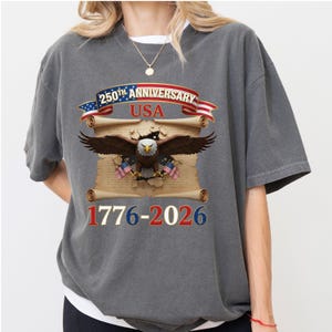 May include: Gray t-shirt featuring a patriotic design for the 250th Anniversary of the USA. The design includes an eagle, American flags, and the years 1776-2026. The text "250th Anniversary USA" is displayed on a banner.