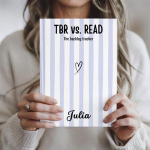 May include: A rectangular book with a white and light purple vertical striped cover. The text reads TBR vs. READ, The backlog tracker, and Julia. A small black hand-drawn heart is centered on the cover. The book is held in a neutral position.