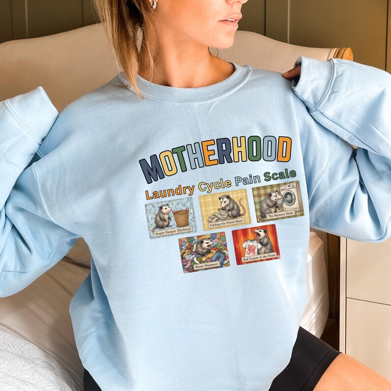 May include: Light blue sweatshirt with the text "MOTHERHOOD Laundry Cycle Pain Scale". The design features cartoon opossums in various laundry-related scenarios. The sweatshirt is a crew neck style.
