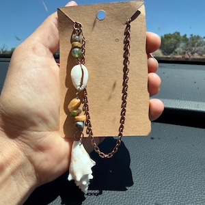 May include: A copper-toned metal chain necklace featuring a series of natural elements including small green and tan stone beads, a white cowrie shell, and a larger white spiral seashell pendant, all displayed on a rectangular brown card.
