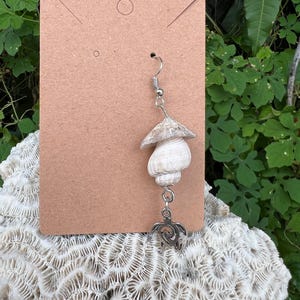 Może przedstawiać: Kolczyk z muszli z grzybowym topem i zawieszką w kształcie żółwia. Kolczyk wykonany jest z naturalnych muszli i metalu w kolorze srebrnym. Kolczyk jest prezentowany na brązowej karcie.