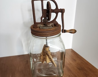 May include: An antique glass butter churn with a rusty metal top and handle. The churn has a wooden paddle inside and the word "PATENT" is visible on the metal frame. The glass jar is clear and sits on a wooden surface.