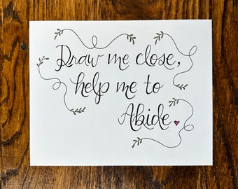 May include: A white card with black calligraphy that reads "Draw me close, help me to abide." The text is surrounded by a black vine with green leaves and a small pink heart.