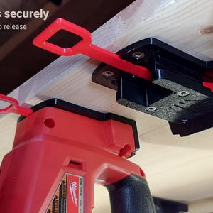 Peut inclure: Support d'outil électrique rouge et noir monté sur une surface en bois. Le support est doté d'une tirette rouge pour une libération facile. Le texte indique "Locks securely, Pull tab to release". L'outil est rouge et noir.