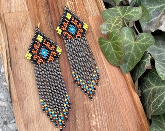 May include: A pair of long, beaded earrings with a geometric pattern in black, orange, yellow, and blue. The earrings are hanging from a wooden surface.