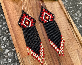 Puede incluir: Un par de aretes largos de cuentas negras con patrones geométricos rojos y dorados. Los aretes tienen una franja de cuentas negras, rojas y blancas.