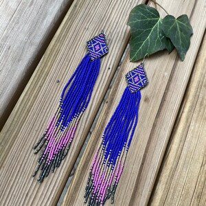 Puede incluir: Un par de pendientes largos de cuentas con un patrón geométrico en rosa, azul y plateado. Los pendientes cuelgan de una superficie de madera.