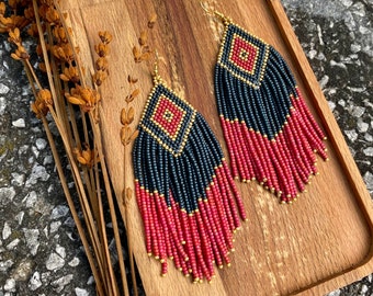 May include: A pair of long, beaded earrings with a geometric pattern in black, red, and gold. The earrings are hanging from a wooden tray.