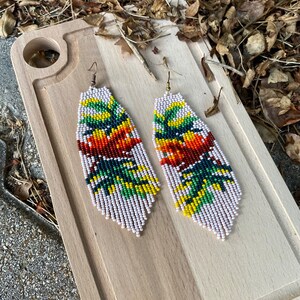 Puede incluir: Un par de aretes largos de cuentas con una base blanca y diseños florales coloridos. Los aretes presentan una mezcla de cuentas rojas, naranjas, amarillas, verdes y azules.