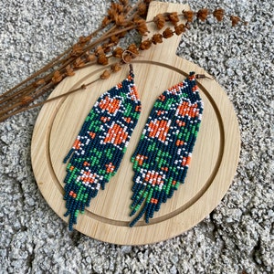 May include: A pair of long, beaded earrings with a geometric pattern in shades of green, orange, white, and black. The earrings are hanging on a wooden surface.