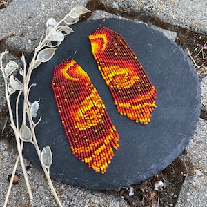 May include: A pair of long, beaded earrings with a red, orange, and yellow color scheme. The beads are arranged in a pattern that resembles a sunset or a flame.