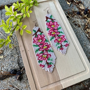 May include: A pair of long, beaded earrings featuring pink flowers and green leaves. The earrings are displayed on a wooden surface.