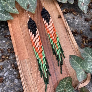 May include: A pair of long, beaded earrings with a black, green, orange, and peach color scheme. The earrings are hanging from a wooden surface.