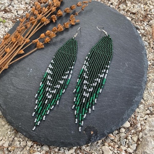May include: A pair of beaded earrings with a fringe design. The earrings are primarily dark green with white and silver accents. The earrings are displayed on a dark gray, round stone surface.