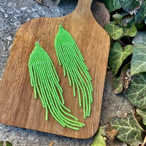 Op de afbeelding: Een paar lange, groene kralen oorbellen met een franje ontwerp. De oorbellen hangen op een houten oppervlak.