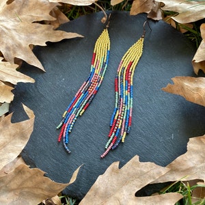 May include: A pair of long, colorful beaded earrings with a yellow, red, blue, and green color scheme. The earrings are hanging from a dark gray surface.