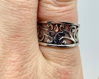 Puede incluir: Anillo plateado con un diseño calado con motivos florales y de volutas. El anillo se lleva en un dedo con esmalte de uñas azul. El diseño del anillo es visible.