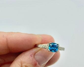 May include: A silver ring with a striking square, blue gemstone at its centre. The band is embellished with smaller, clear gemstones. The ring is set against a plain white backdrop, accentuating its elegant design.