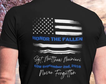 May include: Black t-shirt featuring a distressed American flag design with the text "Honor the Fallen." Below the flag are the words "Sgt. Matthew Mairieri EOW September 2nd, 2018 Never Forgotten."