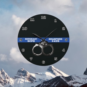 May include: Black clock with white numbers and hands. The clock face has a blue and white police ribbon with the text "SERGEANT SCPD" and "BRANFORD #439".  A pair of silver handcuffs are centered on the clock face.