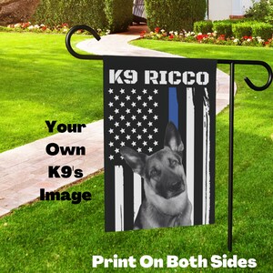 May include: A black and white garden flag with a German Shepherd dog and the text "K9 Ricco". The flag has a blue line and white stars on a black background. The flag is printed on both sides.