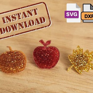 May include: Three glitter-covered cutouts: a red apple, a gold pumpkin, and a gold leaf. The cutouts are on a wooden surface. The text "INSTANT DOWNLOAD" is visible in a red stamp above the cutouts. The text "SVG" and "DXF" are visible in purple and orange boxes, respectively, in the upper right corner of the image.